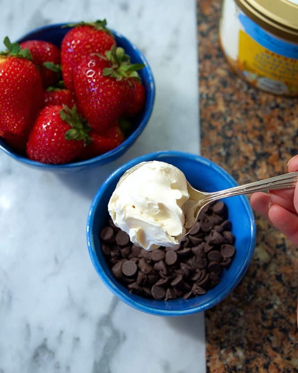 A white woman's hand is holding a silver spoon with a white creamy substance close to a blue bowl filled with dark brown chocolate chips. Behind it, there is another blue bowl full of bright red strawberries with green leaves. Both bowls are placed on a white marbled textured surface with a brown and white speckled pattern. Part of a container with a yellow and blue label is visible on the right side. photo taken with an iphone --ar 4:5 --v 7