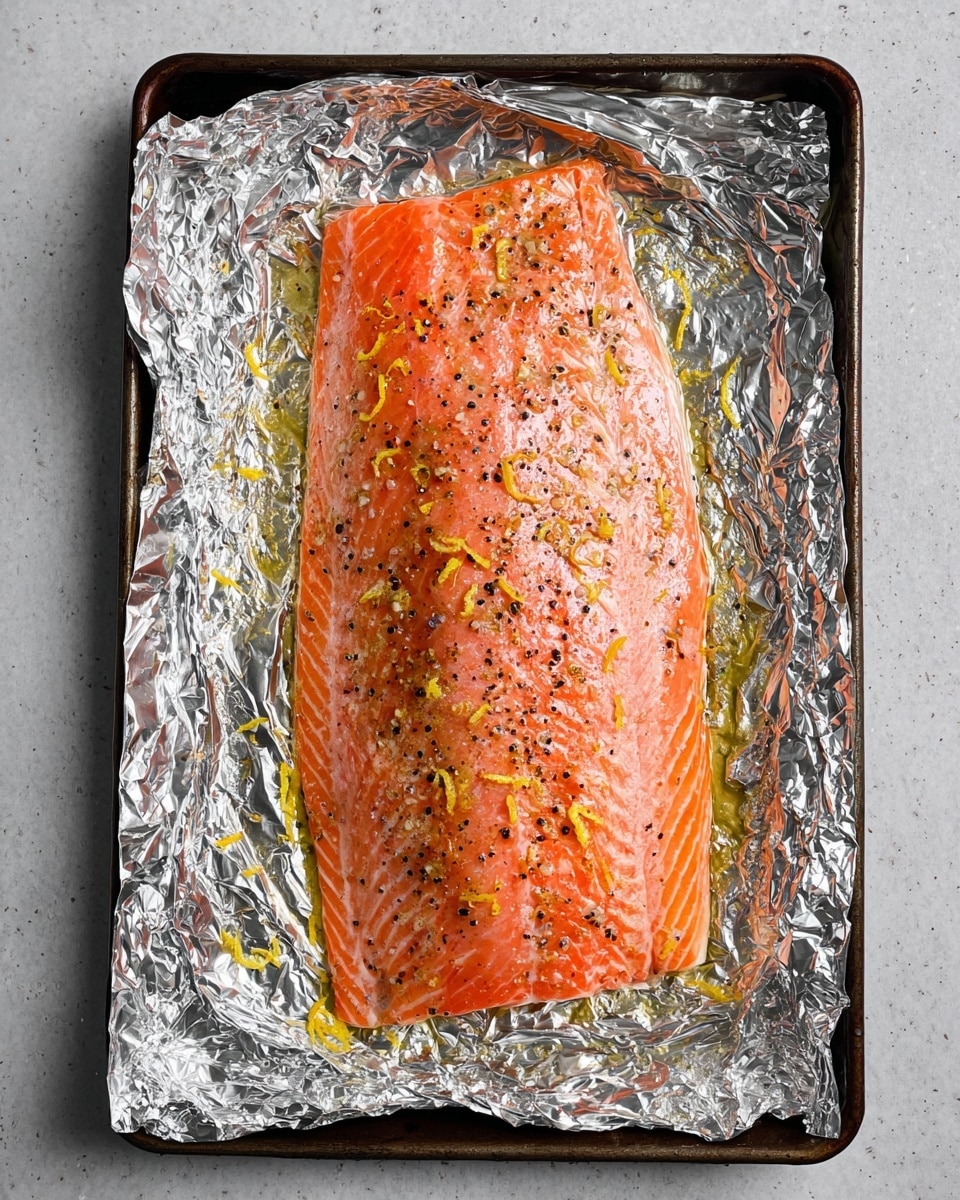 A large piece of cooked salmon with a bright pink-orange color lies flat on crumpled shiny silver aluminum foil that covers a dark baking tray. The salmon is sprinkled with small black pepper flakes and thin yellow lemon zest strips. The surface underneath the tray is white marbled texture. photo taken with an iphone --ar 4:5 --v 7