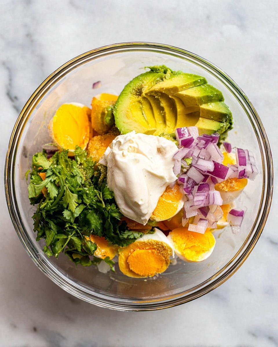 In a clear glass bowl on a white marbled surface, there are several layers of ingredients. The bottom layer shows chunks of bright orange yolks that are slightly soft, next to a half avocado with green and yellow flesh. On top of this, scattered pieces of chopped red onions add a touch of purple. There is also a small bunch of fresh green cilantro on one side. At the top, a dollop of white creamy mayonnaise sits above the avocado and onions. The bowl's clear edges and the smooth textures of the ingredients give a fresh and colorful look. photo taken with an iphone --ar 4:5 --v 7