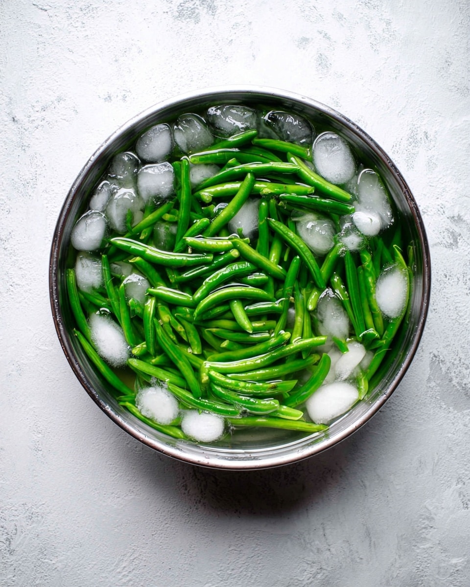 A round metal bowl filled with bright green beans submerged in clear water with many small ice cubes floating around, the green beans are evenly spread and partially overlapped, showing a shiny, fresh texture. The bowl sits on a surface with a white marbled texture, creating a clean and simple background. photo taken with an iphone --ar 4:5 --v 7