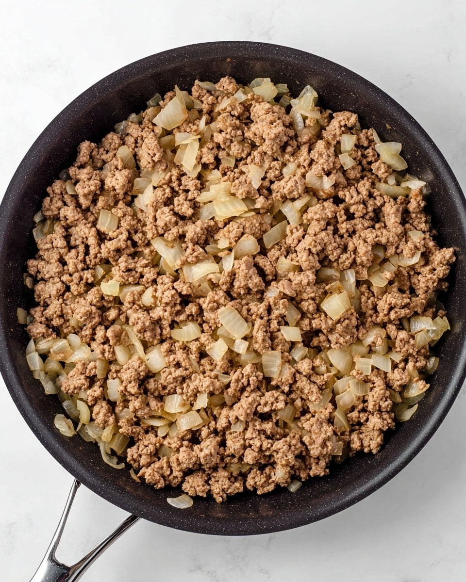 A close-up top view of browned ground meat mixed with cooked diced onions in a black frying pan with a silver handle, placed on a white marbled surface. The cooked meat is light brown with rough, crumbly texture, and the onions are translucent and soft, scattered evenly throughout the dish. The pan is centered in the image, filling most of the frame. Photo taken with an iphone --ar 4:5 --v 7