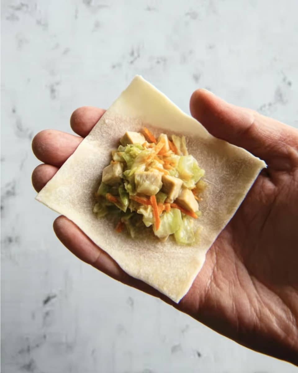 A woman's hand holds a square piece of thin, light beige dough with a slightly translucent and soft texture. On top, centered, there is a small pile of chopped filling consisting of light yellow tofu pieces mixed with small, bright green cabbage leaves and thin orange carrot strips. The background is a white marbled texture. photo taken with an iphone --ar 4:5 --v 7