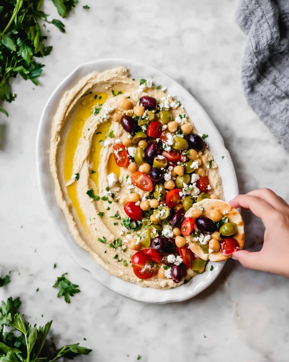 A white oval plate holds a thick layer of smooth beige hummus spread evenly, drizzled with light golden olive oil streaks on the left side. On the right side, a colorful mix of halved red cherry tomatoes, whole green and black olives, small white cheese crumbles, chickpeas, and green parsley leaves adds texture and color contrast. A woman's hand is reaching from the right holding a piece of round pita bread topped with hummus, chickpeas, a black olive, and a cherry tomato piece. The plate sits on a white marbled surface with some blurred green herbs and a gray cloth in the background. Photo taken with an iphone --ar 4:5 --v 7