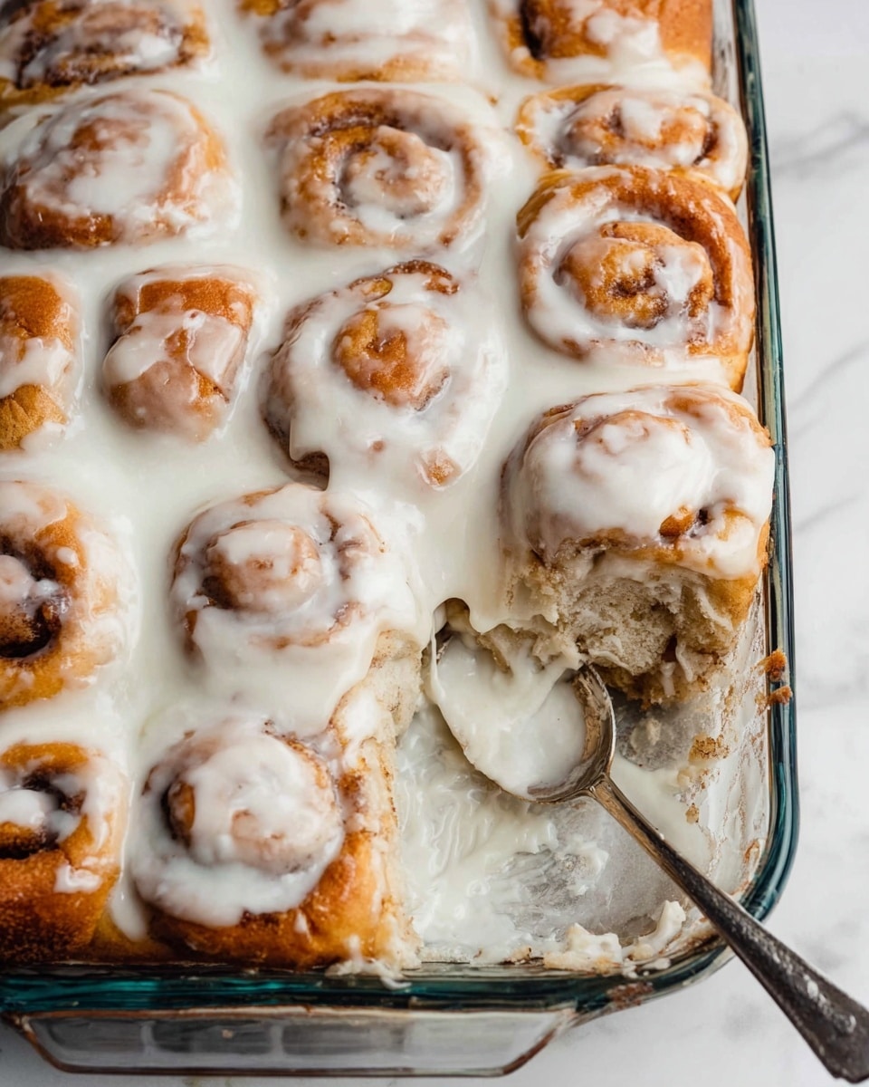The image shows a glass baking dish filled with about three layers of round cinnamon rolls tightly packed together. The cinnamon rolls have a golden-brown color with visible swirls of cinnamon, and they are generously covered with thick white icing that drips down the sides and fills some gaps in between. A silver spoon is partially submerged in the icing at the lower right corner of the dish. The dish is placed on a surface with a white marbled texture. photo taken with an iphone --ar 4:5 --v 7