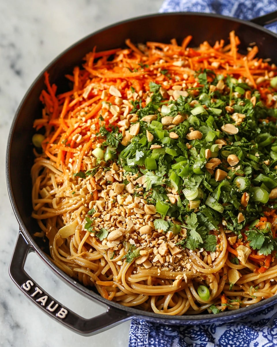 In a black cooking pan, a bed of long light brown noodles fills the bottom layer. On top, there is a mix of shredded bright orange carrots, chopped green onions, fresh green cilantro, light brown crushed peanuts, and small white sesame seeds evenly spread over the noodles. The ingredients create a colorful and fresh look with a mix of textures. The pan handle shows the word