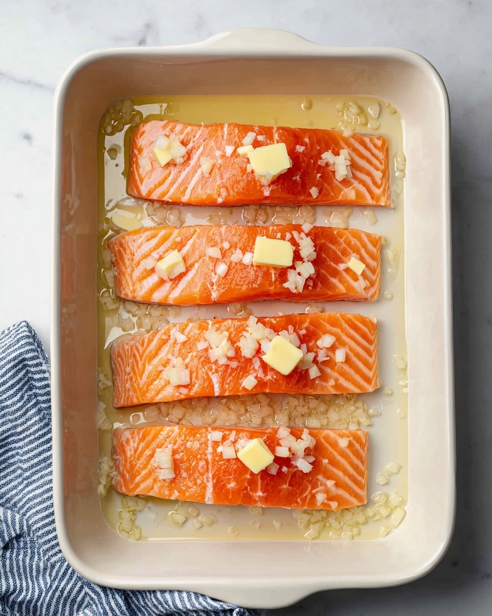 A white rectangular baking dish holds four pieces of raw salmon fillets arranged in a single layer from top to bottom. Each fillet is bright orange with light stripes of white fat. Small pieces of chopped shallots are sprinkled over the salmon and the bottom of the dish. There are several small yellowish butter slices spaced evenly on and between the fillets. The dish contains a light liquid marinade collecting at the bottom around the salmon. The dish is placed on a white marbled surface with a striped blue and white cloth partially visible on the lower left corner. Photo taken with an iphone --ar 4:5 --v 7