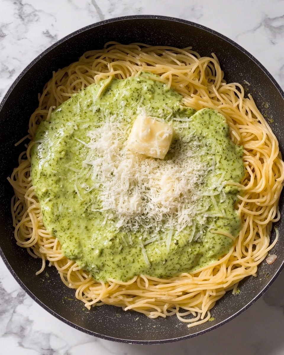 A black pan holds three layers of food: the bottom layer consists of lightly yellow cooked spaghetti noodles spread evenly across the pan, the middle layer is a thick, creamy green sauce with specks of darker green herbs, poured mostly towards the center covering some noodles but leaving the edges visible, and the top layer includes a small pat of pale yellow butter melting in the sauce along with a generous sprinkle of finely grated white cheese scattered around the butter and on the green sauce. The pan sits on a white marbled surface. photo taken with an iphone --ar 4:5 --v 7
