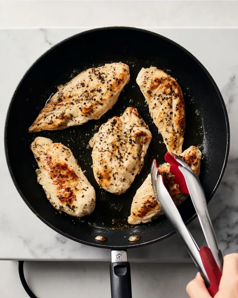 A black pan on a white marbled surface holds four cooked chicken pieces with light brown sear marks and black pepper on top. The chicken pieces vary in size and shape, placed with three pieces near the top and one longer piece near the bottom. A woman's hand with silver tongs that have red silicone tips is holding the piece on the right side in the pan. Photo taken with an iphone --ar 4:5 --v 7