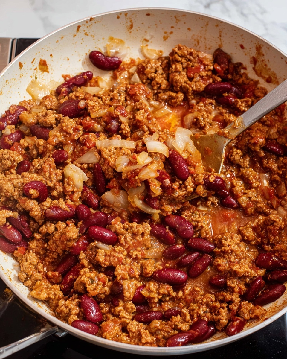 A close-up showing a frying pan with cooked ground meat mixed with dark red kidney beans and finely chopped translucent white onions. The mixture is coated in a thick reddish-brown sauce, with the spoon lifting a portion of the mix in the center. The pan has a white inside and is set on a stovetop with a black surface around it, and the background is a white marbled texture. photo taken with an iphone --ar 4:5 --v 7