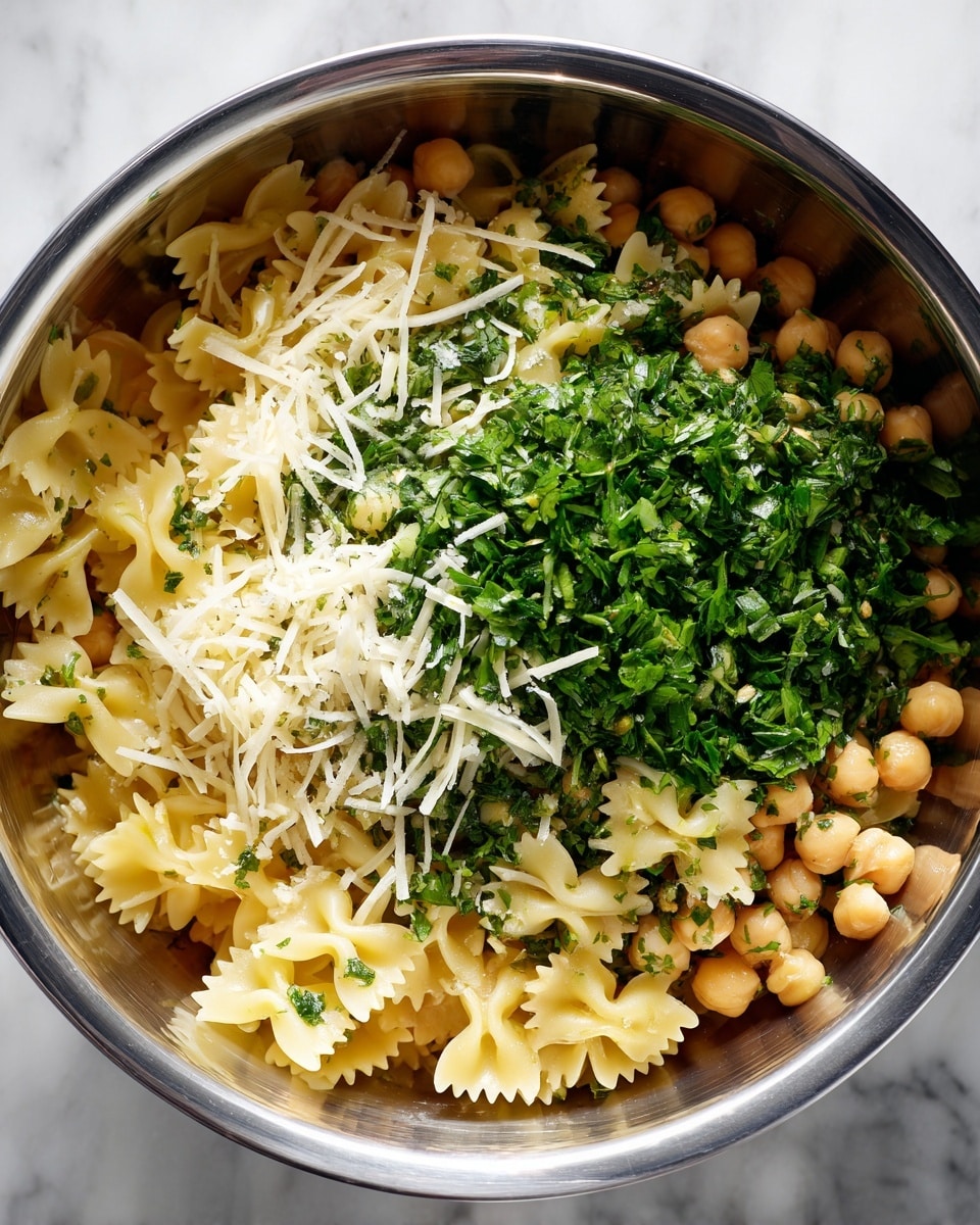The image shows a close-up of a bowl with three main layers. The bottom layer is light yellow pasta shaped like small bow ties, mixed with pale beige chickpeas scattered evenly. On top of this is a vibrant green layer of chopped fresh herbs, likely parsley, spread across. The top layer consists of a light sprinkling of finely shredded white cheese, concentrated more on one side. The bowl itself is metallic and shiny, with a hint of reflection, all set on a surface with a white marbled texture. Photo taken with an iphone --ar 4:5 --v 7