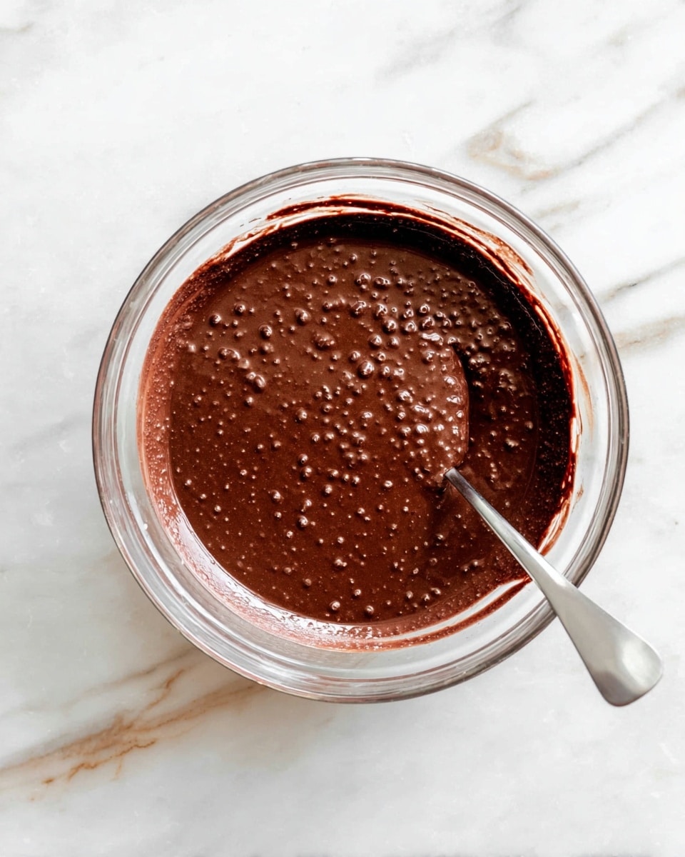 A clear glass bowl is filled with a thick, dark brown chocolate mixture that has small air bubbles all over the surface, giving it a slightly textured look. A metal spoon sticks out from the right side, partly submerged in the mixture and resting near the center. The bowl sits on a white marbled surface with light gray and beige veins. The edge of the bowl has some chocolate smudges. Photo taken with an iphone --ar 4:5 --v 7