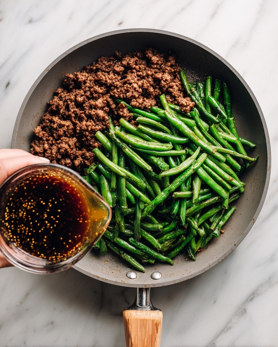 A gray frying pan with wooden handle sits on a white marbled surface, containing a layer of cooked dark brown ground meat spread evenly across the bottom. On top, in the middle, there is a pile of bright green chopped green beans with a shiny surface, showing freshness and slight cooking. At the bottom center of the image, a woman's hand holds a clear glass measuring cup filled with dark brown sauce with visible mustard seeds and a slightly thick texture, about to pour it over the green beans and meat. Photo taken with an iphone --ar 4:5 --v 7
