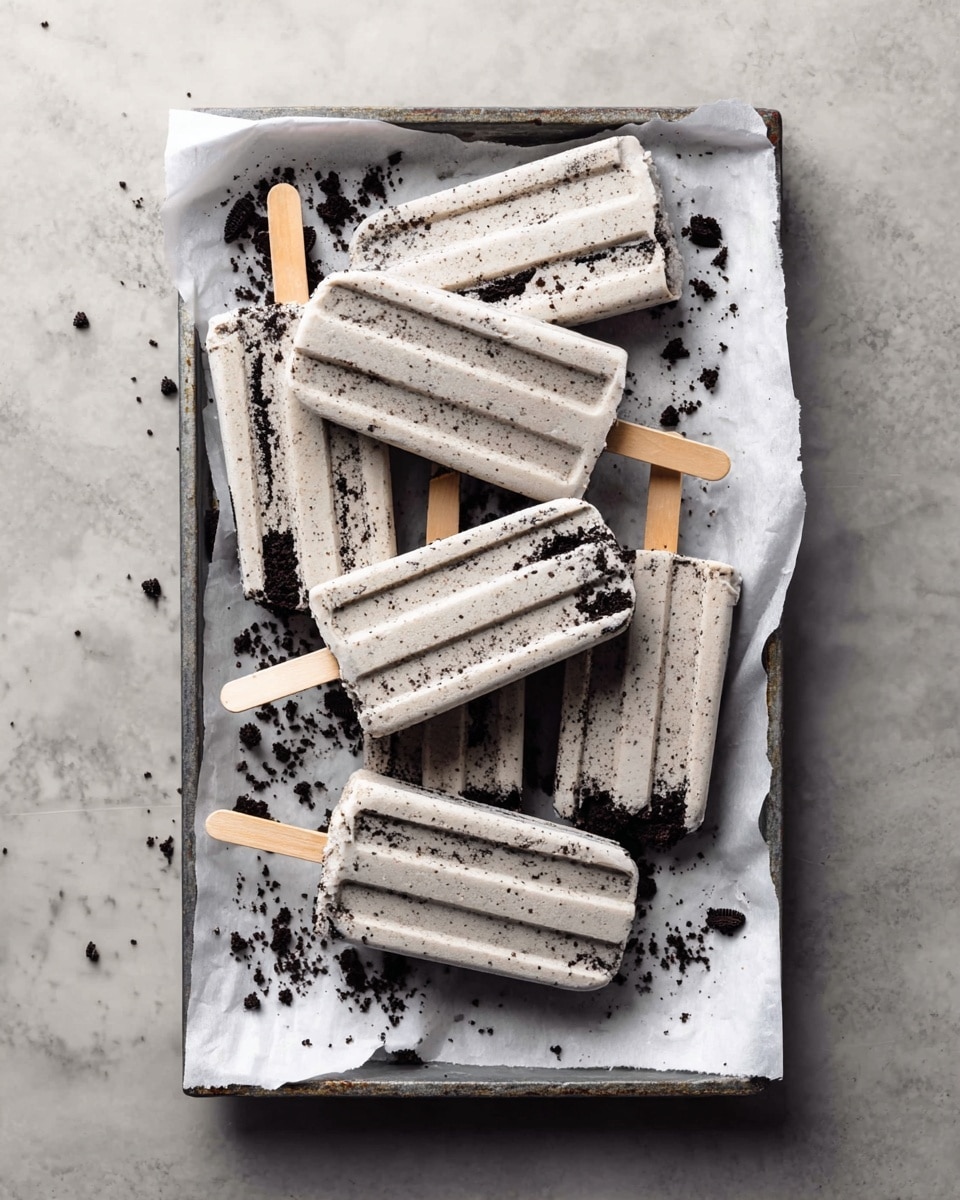 A tray lined with white parchment paper sits on a white marbled surface, holding seven light gray popsicles with dark cookie crumbs mixed inside. The popsicles have a slightly creamy texture with crumb bits unevenly spread throughout and distinct ridges on the top surface, each popsicle having a light wood stick inserted at the bottom. Around the popsicles are small crumbled pieces of the dark cookies, adding a scattered effect. The scene has a neutral, soft light that highlights the texture and cool tone of the popsicles and the contrast with the darker cookie crumbs. Photo taken with an iphone --ar 4:5 --v 7