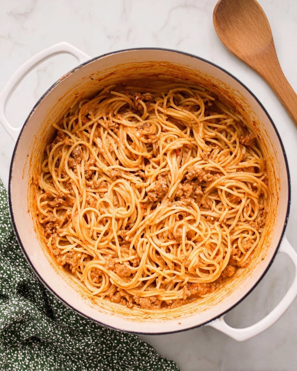 A white large pot filled with long, thin spaghetti mixed in a light red creamy sauce, with small pieces of light brown cooked meat scattered evenly throughout. The inside sides of the pot have sauce smudges showing a rich texture. On the right, there is a wooden spoon resting on a white marbled surface, and on the left, part of a green cloth with white patterns is visible. The overall color scheme is soft reds, light browns, and the clean white of the pot and surface. Photo taken with an iphone --ar 4:5 --v 7