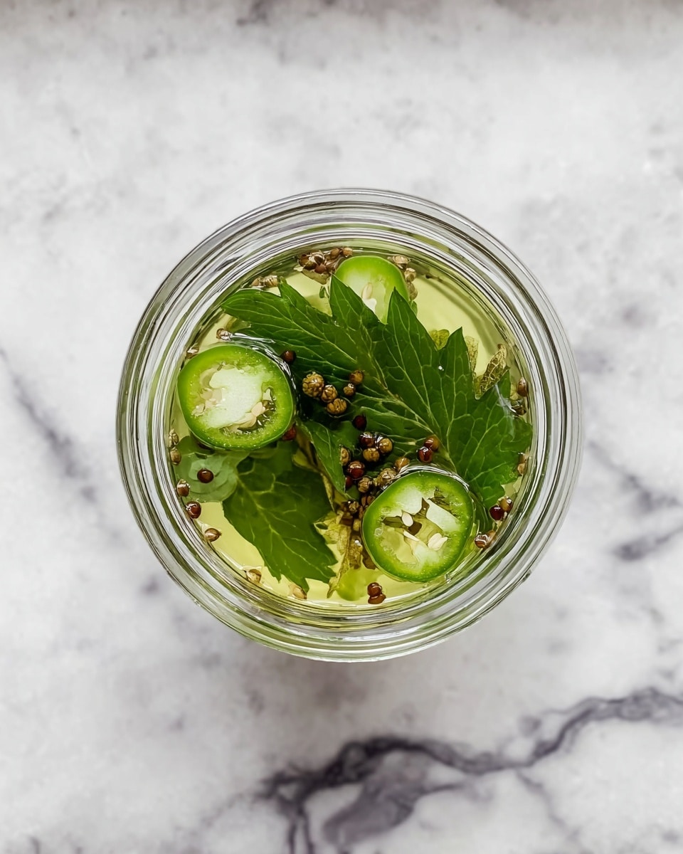 A clear glass jar seen from above with three main layers inside: at the bottom, pale yellow-green liquid, in the middle several dark green leaves that are fresh and crisp, and on top, scattered sliced round green jalapeños and small brown mustard seeds floating on the liquid. The jar is placed on a white marbled surface showing gray and white veins. photo taken with an iphone --ar 4:5 --v 7