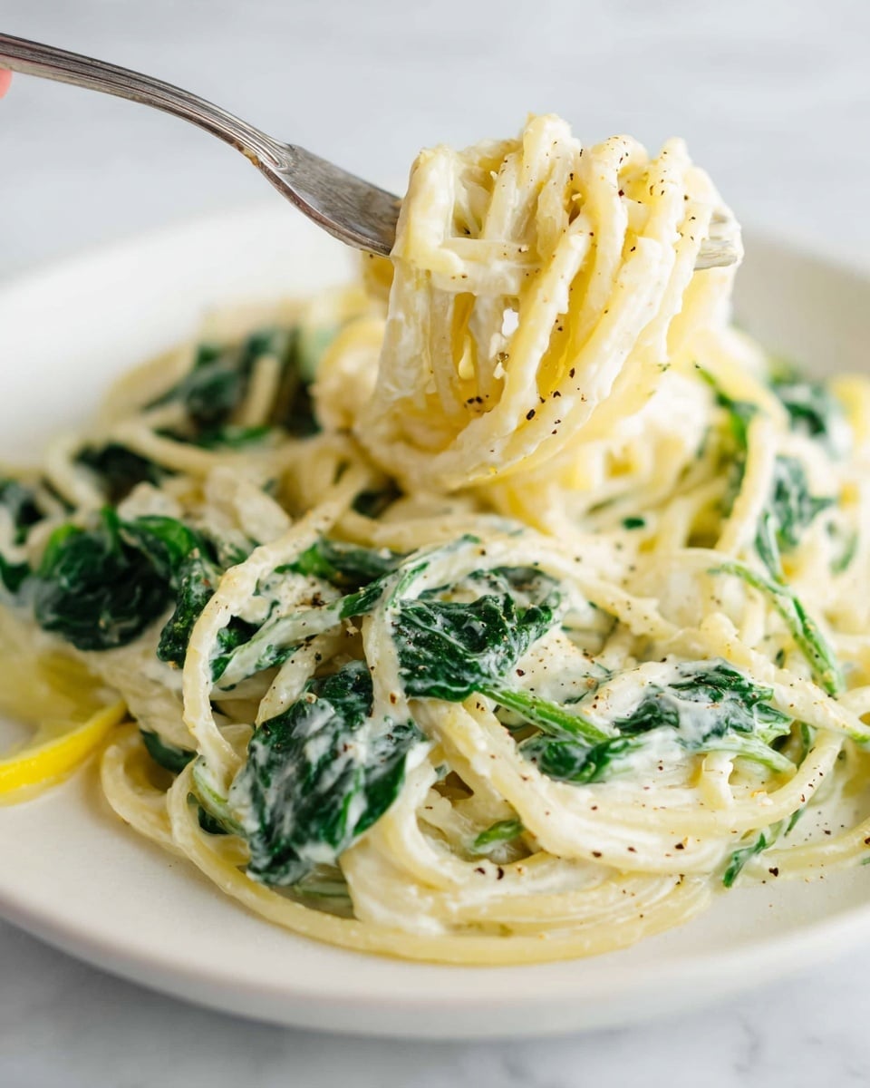 A close-up of creamy pasta served on a white plate with a white marbled surface underneath. The dish has long spaghetti noodles coated in a smooth, light cream sauce, mixed with wilted green spinach leaves. The top layer shows the noodles twirled around a fork (held by a woman's hand), revealing the soft texture and a slight dusting of black pepper. The colors are mainly creamy white from the sauce and bright green from the spinach, with a few small touches of yellow lemon slices visible under the noodles. The scene is simple and clean with a focus on the creamy texture and fresh greens photo taken with an iphone --ar 4:5 --v 7
