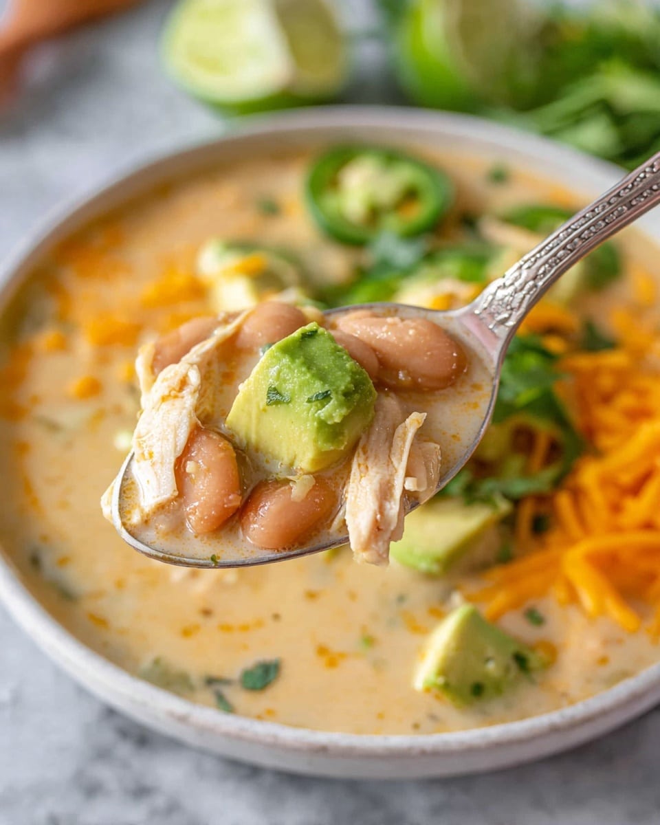 A close-up of a spoon holding a light tan creamy soup with soft light pink beans, shredded light beige chicken, and a small green avocado chunk; in the background, the soup is in a white bowl filled with chunks of green avocado, slices of green jalapeño, orange shredded cheese around the edges, two lime wedges, and fresh green herbs, all on a white marbled surface. photo taken with an iphone --ar 4:5 --v 7