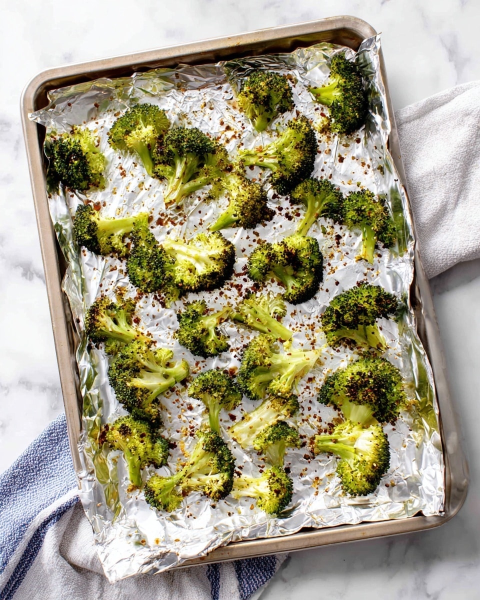 A baking tray lined with crinkled silver foil holds many pieces of roasted broccoli scattered across the surface. The broccoli pieces are bright green with slightly darkened, crispy edges showing light charring. Tiny bits of browned seasoning are sprinkled unevenly over the foil and broccoli. The tray rests on a white marbled surface with a white and blue cloth partially visible underneath one corner of the tray. photo taken with an iphone --ar 4:5 --v 7