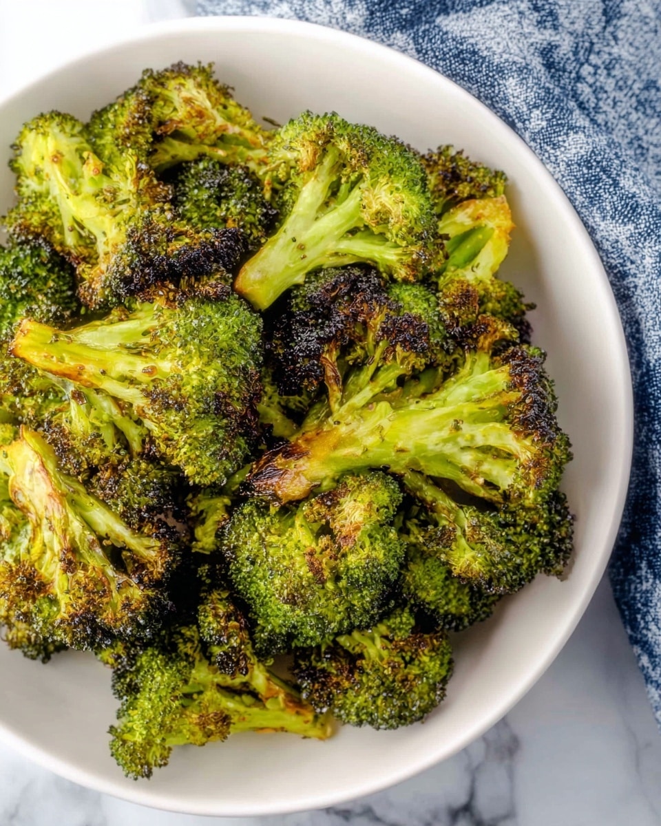 A white bowl filled with roasted green broccoli florets that have crisp, dark brown edges showing slight charring, giving a textured mix of bright green and caramelized colors. The broccoli pieces are layered on top of each other, showing the woody stems and tightly packed green buds. The bowl sits on a white marbled surface with a blue and white cloth partially visible on one side. Photo taken with an iphone --ar 4:5 --v 7