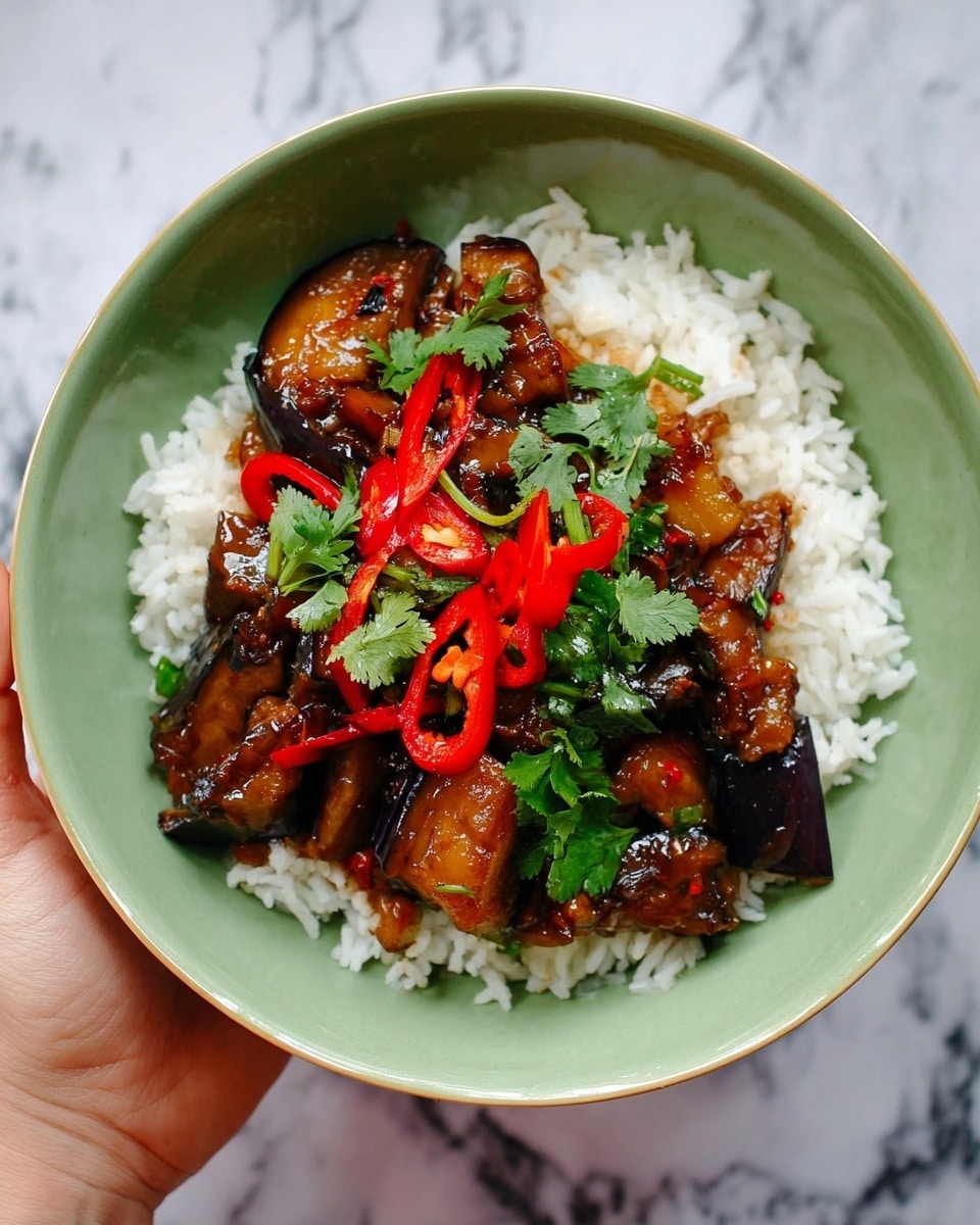 A white bowl filled with a base layer of fluffy white rice, topped with a glossy dark brown stir-fry mix of eggplant pieces coated in sauce. On top, there are sliced bright red chili peppers and green herbs, including fresh cilantro leaves, adding color contrast. The bowl rests on a white marbled surface with a woman's hand holding it from behind, slightly entering the frame from the top. Photo taken with an iphone --ar 4:5 --v 7