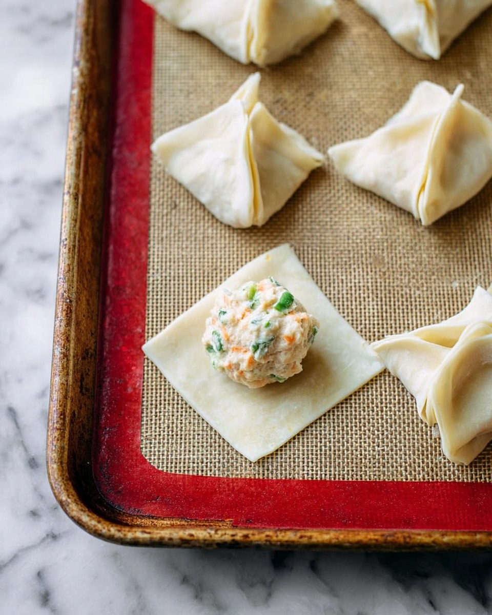 The image shows a baking tray with a red frame and a textured mat inside. On the mat, there are several folded dumplings with a light beige dough, each folded into a small, square-shaped packet with folded corners. One dumpling wrapper lies flat with a round ball of a creamy, chunky filling that has bits of green herbs and small orange pieces, placed in the center. The dumplings have a soft, slightly shiny texture, and the scene is set on a white marbled surface. Photo taken with an iphone --ar 4:5 --v 7