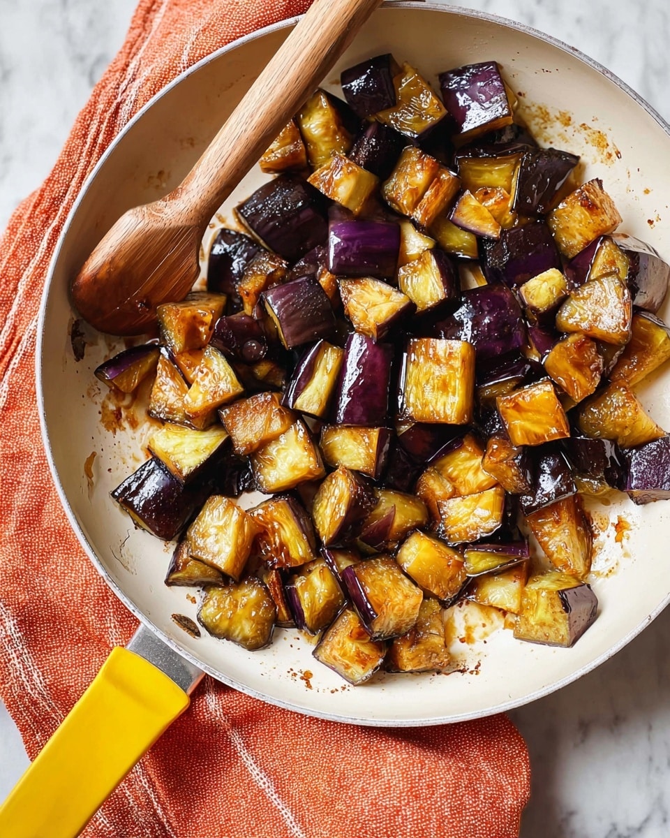 A close-up view of a white pan filled with cooked eggplant pieces, about one layer thick, showing dark purple skin and golden-brown, soft inside, some pieces have a light shiny texture from cooking oil. The pan has a yellow handle with a wooden end, resting on an orange textured cloth, all on a white marbled surface. photo taken with an iphone --ar 4:5 --v 7