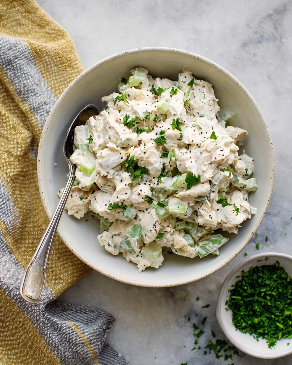 A white bowl filled with a creamy chicken salad showing three main layers: chunky pieces of white chicken mixed with diced light green celery, all coated in a thick, creamy white sauce with visible black pepper and green herbs scattered throughout. On top, small finely chopped bright green parsley is sprinkled evenly. A large silver spoon rests inside the bowl on the left side. To the right, a small white bowl holds extra finely chopped parsley on a white marbled surface. Behind the bowl, there is a yellow and gray striped cloth casually placed. Photo taken with an iphone --ar 4:5 --v 7