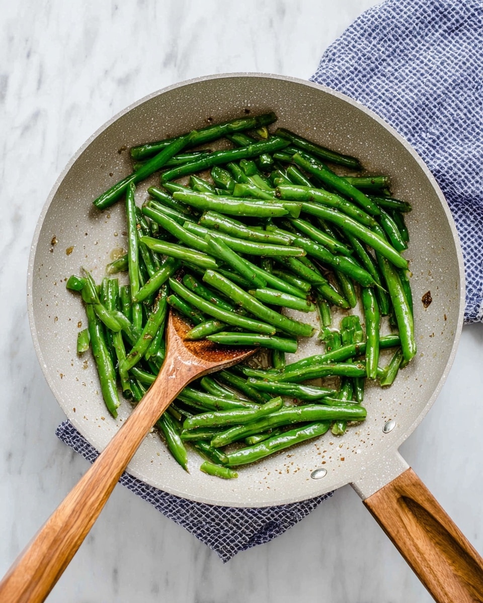 Inside a light gray frying pan with a wooden handle, there are scattered cooked green beans that are shiny with oil, showing some light brown spots from cooking. A wooden spoon with a smooth, warm brown color lies in the pan, partially touching the green beans. The whole scene is set on a white marbled surface with a blue and white checked cloth partially visible under the pan. The colors are fresh and natural, with the glossy green beans contrasting against the soft gray pan and white marbled background. photo taken with an iphone --ar 4:5 --v 7