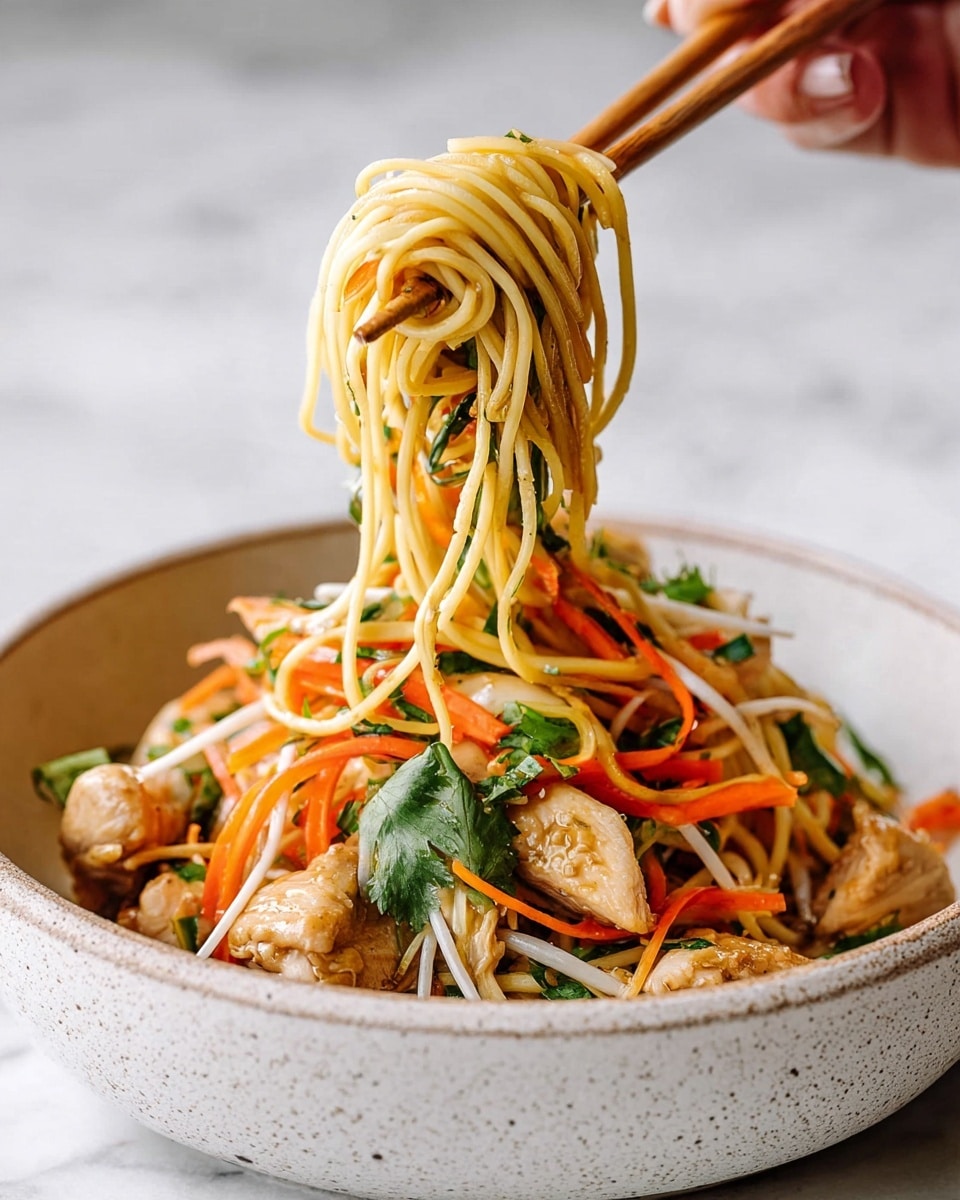 A round white speckled bowl holds a colorful noodle dish. At the bottom, light brown cooked chicken pieces are scattered, mixed with green leafy herbs. On top, there is a thick layer of thin yellow noodles mixed with thin orange carrot strips, white bean sprouts, and leafy greens. A woman's hand with chopsticks lifts a tangle of noodles, showing the mix of bright vegetables and herbs against the lightly sauced noodles. The background and surface have a white marbled texture. photo taken with an iphone --ar 4:5 --v 7