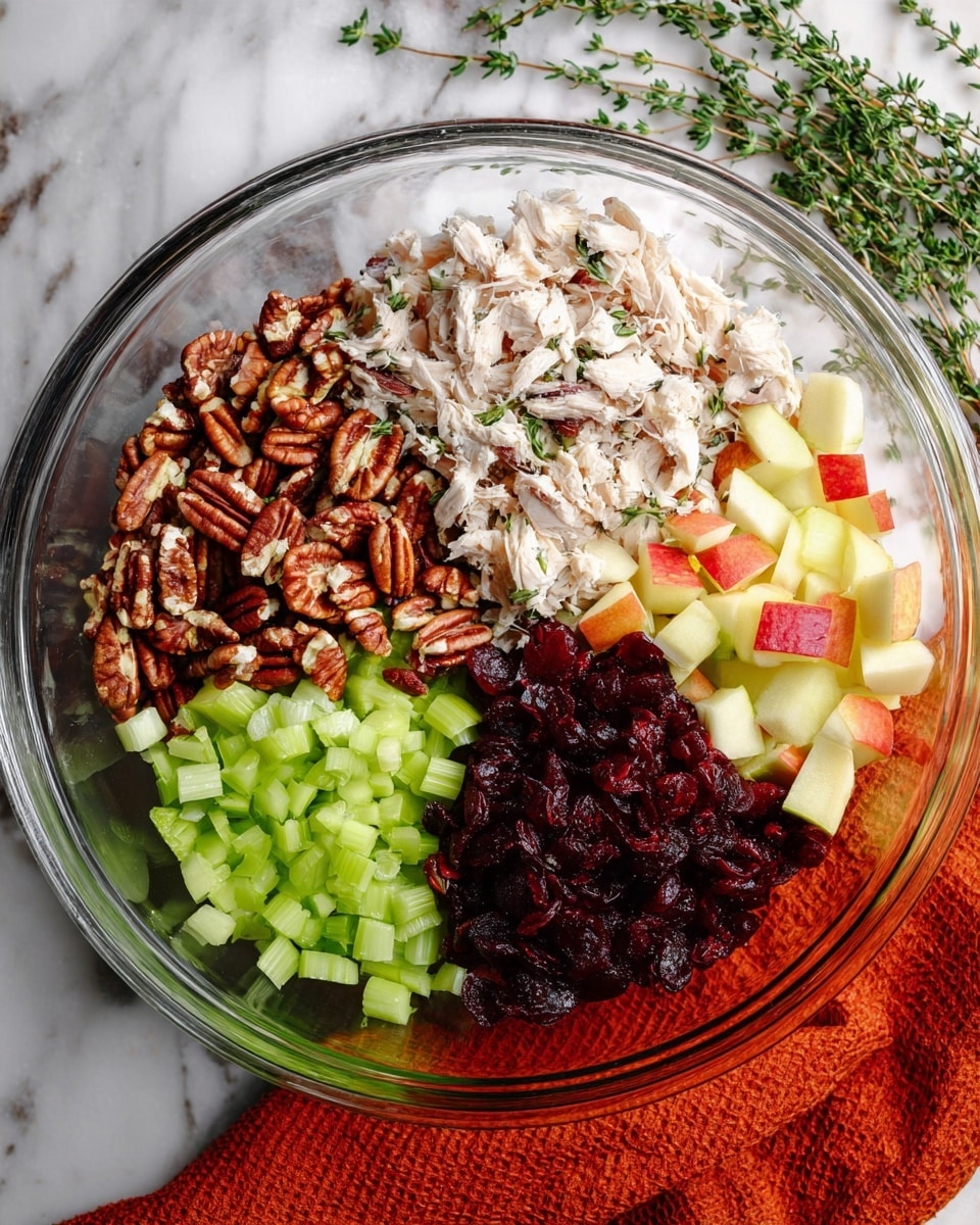 A clear glass bowl on a white marbled surface holds five sections of salad ingredients arranged in a circle: shredded white chicken pieces at the top, chopped pecans with brown and cream colors to the left, diced green celery below the pecans, dark red dried cranberries to the right of celery, and small cubes of light yellow and red apple pieces beside the chicken. Green herb sprigs lie in the background, and an orange cloth is partially visible under the bowl at the bottom right. Photo taken with an iphone --ar 4:5 --v 7