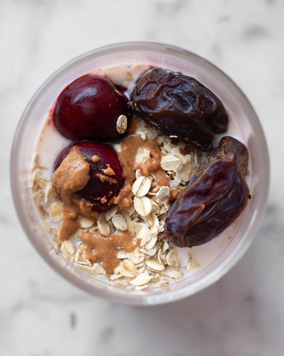 The image shows the inside of a clear glass container with three layers visible. The bottom layer is a pale pinkish-white liquid, likely yogurt or a smoothie base, filling most of the container. On top of this base is a layer of whole dark red cherries, some with a smooth shiny surface and a couple partially coated with a light brown nut butter. Scattered across and around the cherries is a crunchy layer of light beige rolled oats with a rough texture. Near the top edge, there is a large dark brown dried date adding a deep rich color contrast. The overall look is fresh and natural, with soft and rough textures combined, set on a white marbled background. photo taken with an iphone --ar 4:5 --v 7