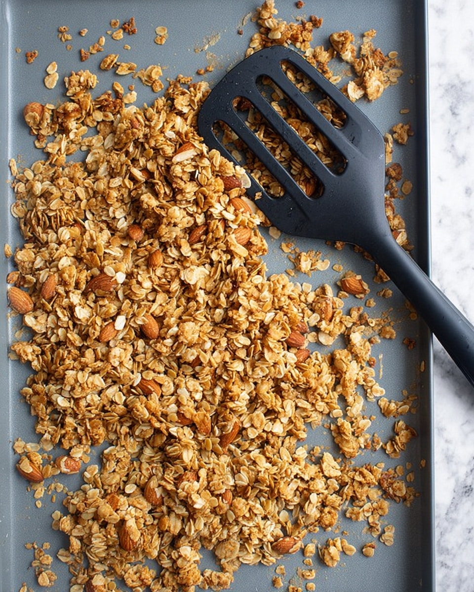 The image shows a single layer of golden brown granola spread across a gray baking pan. The granola mix consists of toasted rolled oats and whole almonds, with a crunchy texture and some small clumps scattered throughout. There is a black slotted spatula resting on the top left side of the pan, partly on the granola. The granola and pan are set on a white marbled surface. photo taken with an iphone --ar 4:5 --v 7