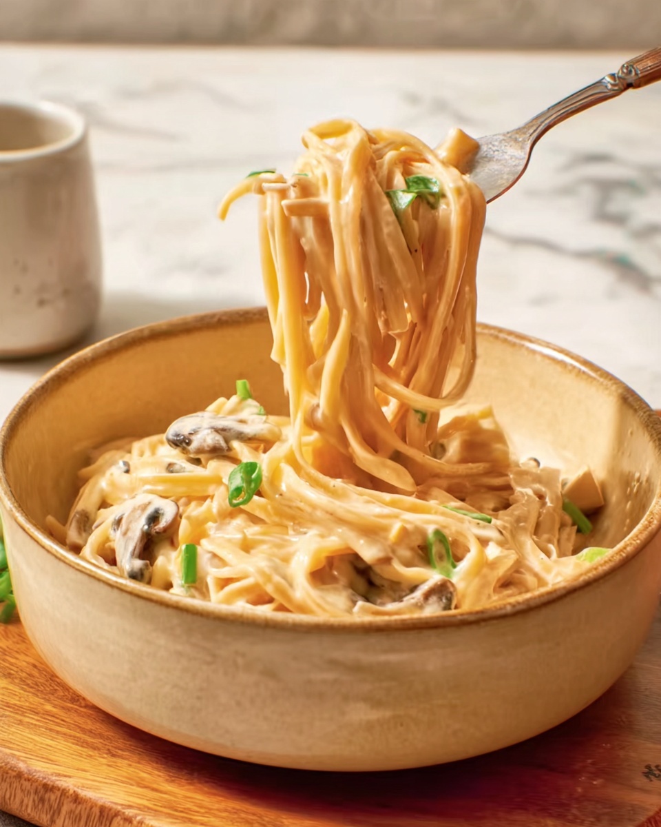 A round, earthy beige bowl holds thick creamy noodles with a light orange sauce, mixed with small cubes of tofu and bits of green herbs scattered on top. A silver fork is lifting a tangled bunch of noodles, showing the creamy texture coating each strand, with some tofu pieces resting beneath the noodles in the bowl. The bowl sits on a wooden board placed on a white marbled surface. In the background, a small white ramekin contains a few green herb pieces, and a soft beige cloth is gently folded nearby. photo taken with an iphone --ar 4:5 --v 7