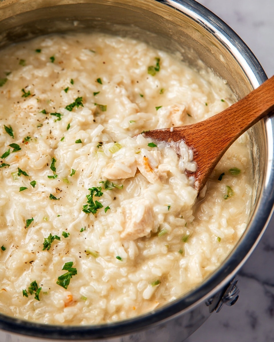 A close-up view of creamy rice dish with small chunks of white meat mixed in, all coated in a smooth, light beige sauce. The dish is garnished with small green herb pieces scattered on top. The creamy rice and meat sit inside a shiny metal pot with a slight reflection visible on the pot's side. A wooden spoon is partly dipped into the dish, showing the textured surface of the rice and sauce. The background surface under the pot is white with a marbled texture photo taken with an iphone --ar 4:5 --v 7