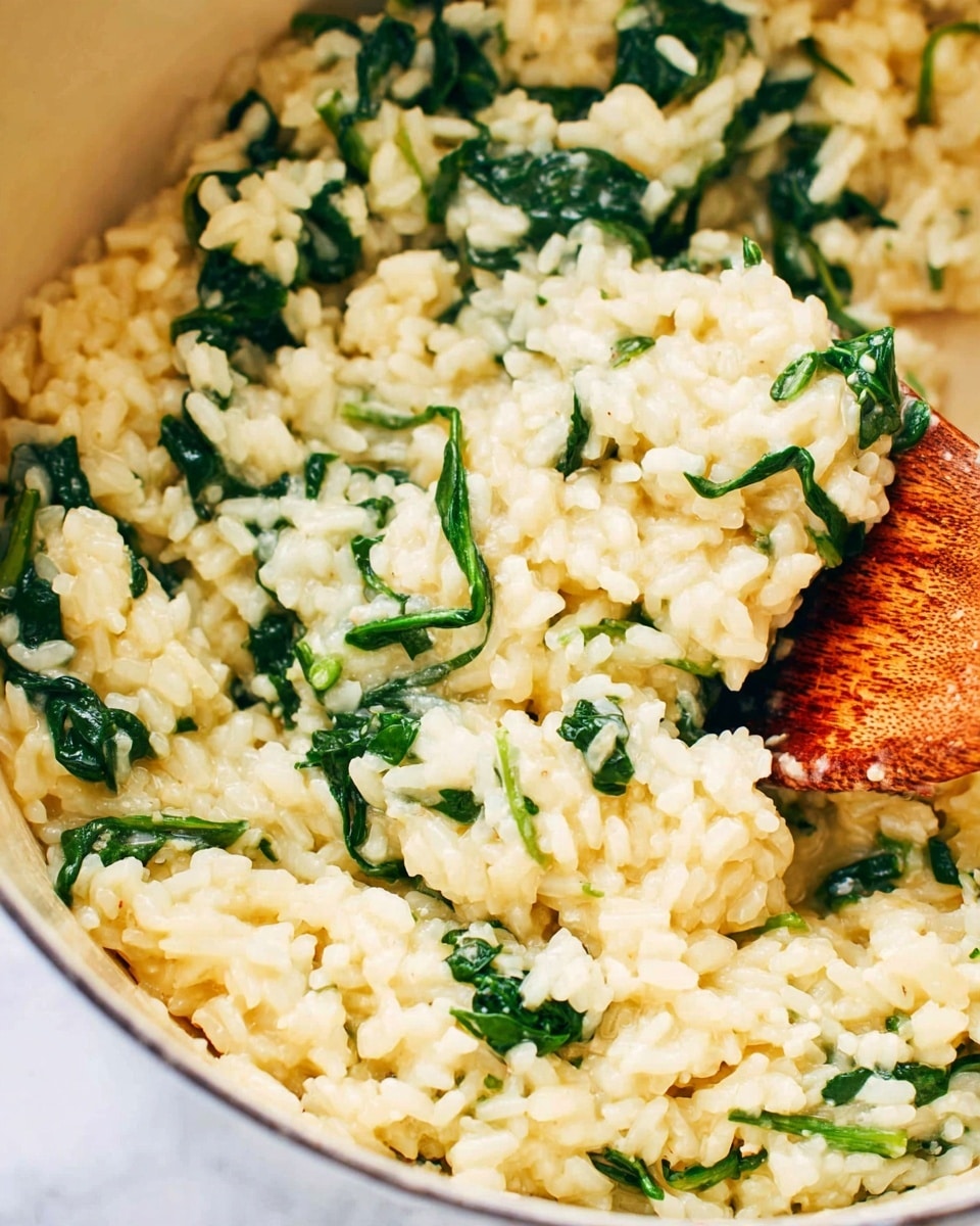 The image shows a close-up of creamy risotto with bright green spinach mixed in. The risotto is light yellow with a soft, moist texture, and the spinach leaves are scattered evenly throughout, adding fresh green color. A wooden spoon is partially visible on the right side, lifting some of the risotto. The background is a white marbled texture, and the dish is inside a white pot. photo taken with an iphone --ar 4:5 --v 7