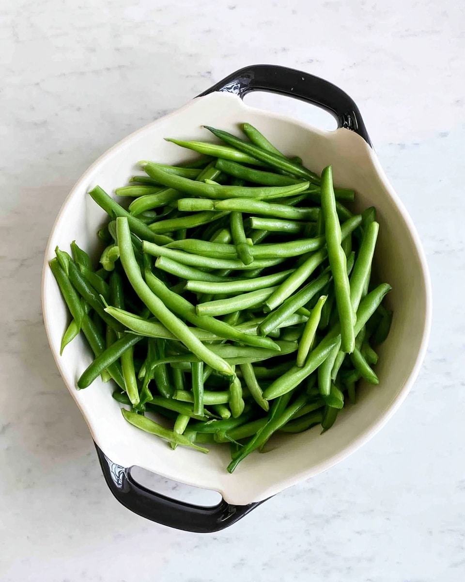 The image shows a bowl full of cooked green beans that are bright and shiny, placed on a soft beige cloth. The bowl is white and shallow, with two wedges of lemon on top adding a pop of yellow color. The green beans are piled in the bowl, with some overlapping and showing their smooth texture and slight wrinkles. The background is a white marbled surface, and a fork is placed beside the bowl. A fresh lemon half is also visible at the bottom left corner of the image, enhancing the fresh feel of the dish. photo taken with an iphone --ar 4:5 --v 7