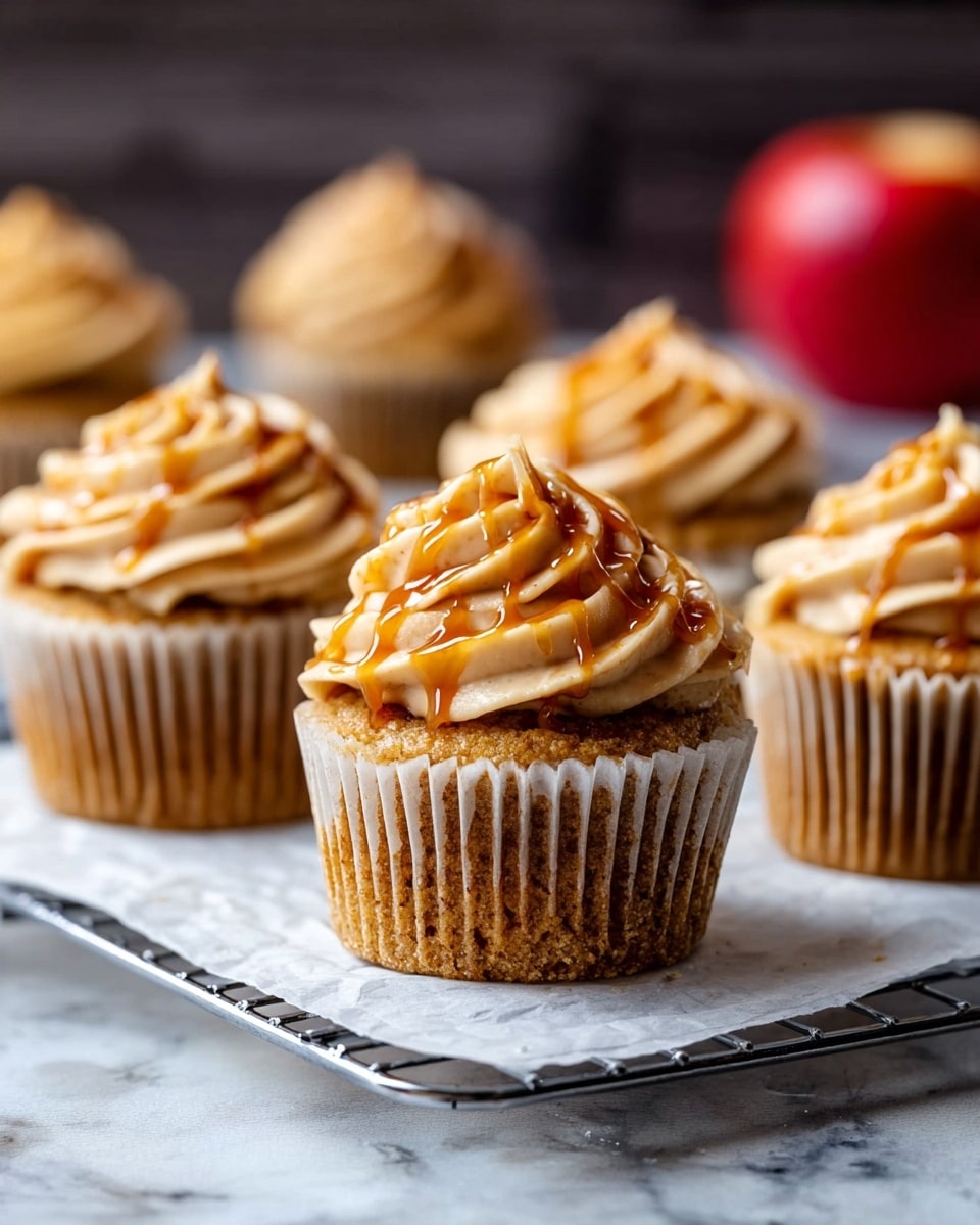 The image shows a close-up of a single cupcake with two main layers: the bottom layer is a brown cupcake base with a textured paper wrapper, and the top layer is a thick swirl of light brown frosting with a smooth, creamy texture, drizzled with shiny caramel sauce. On the very top of the frosting is a thin, triangular slice of red apple with a shiny peel. In the background, there is a similar cupcake slightly out of focus and a whole red apple, all set on a wooden surface with a white marbled textured background. photo taken with an iphone --ar 4:5 --v 7
