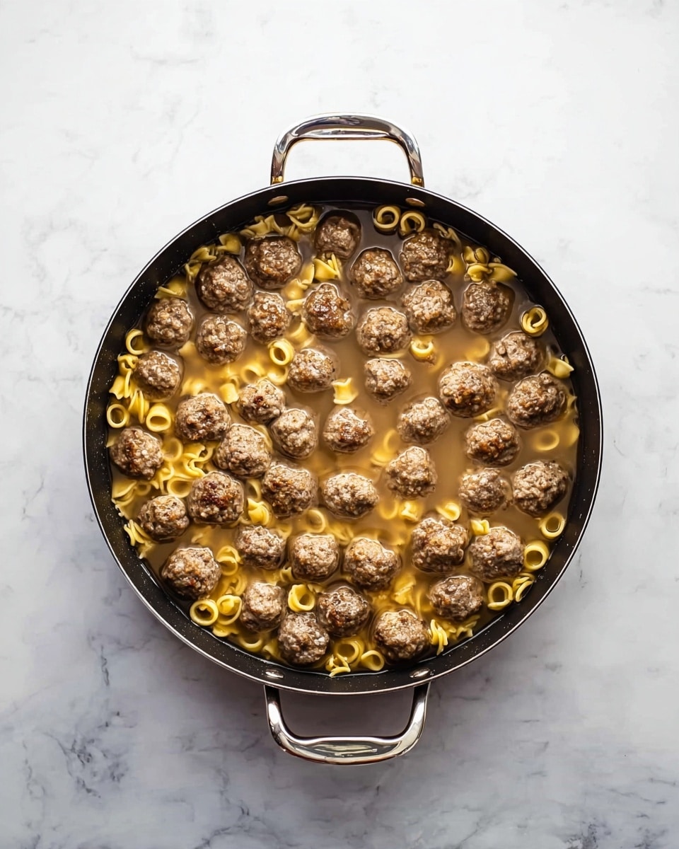 A round black pan with two silver handles holds a mixture of small brown meatballs and yellow ring-shaped pasta pieces partly submerged in a light brown broth. The pan sits on a white marbled surface. The meatballs are evenly spread over the entire surface with pasta scattered mainly around the center and edges. The broth is thin and clear, partially covering the meatballs and pasta. Photo taken with an iphone --ar 4:5 --v 7