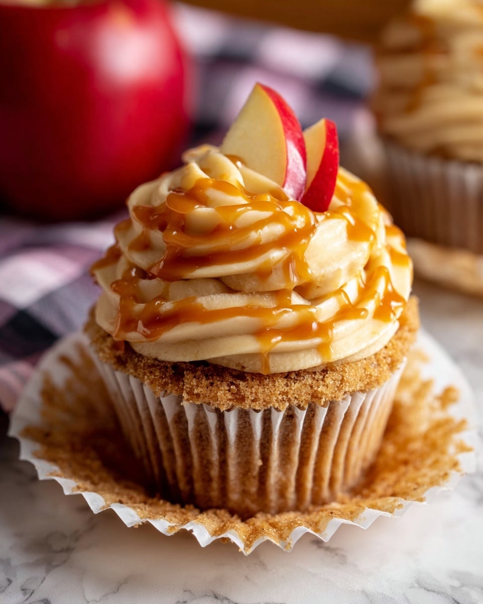 There are five cupcakes on a silver wire rack with white paper underneath, placed on a white marbled surface. Each cupcake has one base layer of golden brown cake with a slightly textured top. On top of each cake is a thick swirl of light tan frosting, with a smooth, creamy texture and a loose spiral shape. A shiny, amber-colored drizzle runs over the frosting on each cupcake, catching the light and adding a glossy effect. In the blurred background, there is a red apple and a dark, out-of-focus backdrop. photo taken with an iphone --ar 4:5 --v 7