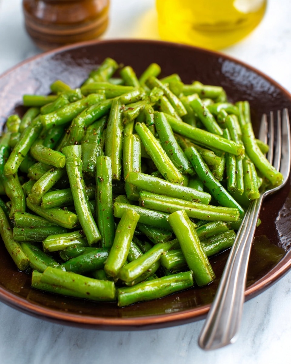 A brown plate filled with chopped cooked green beans with a bit of seasoning, showing their bright green color and smooth texture; in the background, a bottle of olive oil and some blurred objects sit on a white marbled surface, with a silver fork placed at the bottom right corner of the image photo taken with an iphone --ar 4:5 --v 7