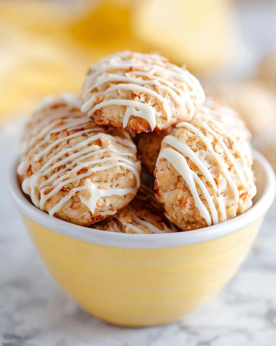 A close-up view of several small golden yellow coconut macaroons lightly toasted on the edges, stacked on top of each other on a white marbled surface with a natural beige woven mat underneath; each macaroon is drizzled with thin white icing lines across the top and sides, showing a rough, shredded coconut texture. In the background, there is a white cup filled with more macaroons and a white glass of milk with a blue and white striped straw. To the right, there is a sliced lemon half visible, all set on a white marbled texture. Photo taken with an iphone --ar 4:5 --v 7