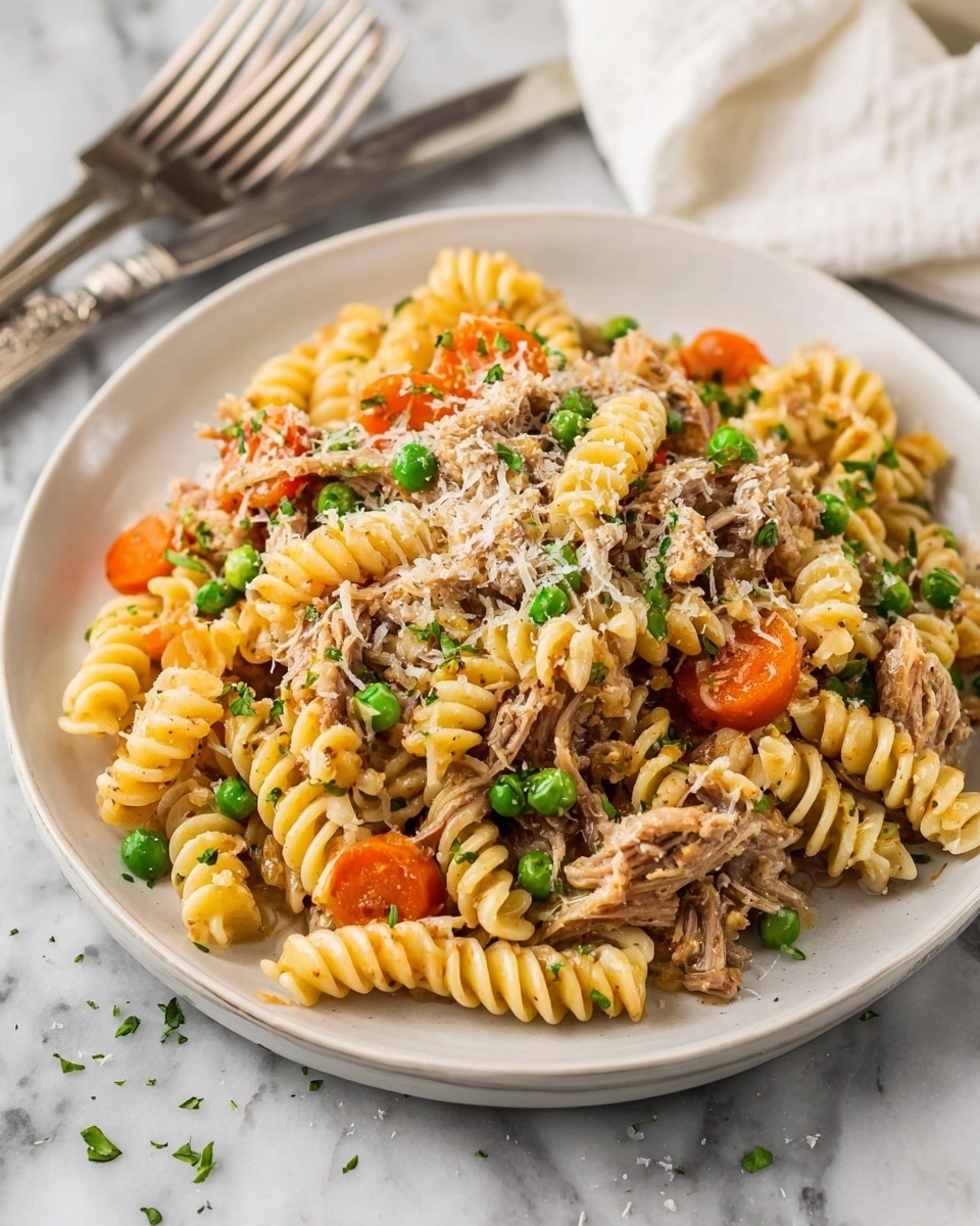 A white plate holds a serving of rotini pasta mixed with shredded light brown meat. Bright orange carrot slices and small green peas are scattered throughout the pasta, adding pops of color. The dish is lightly sprinkled with grated, pale yellow cheese on top. The plate sits on a white marbled surface, with two silver forks and a white cloth in the background. Photo taken with an iphone --ar 4:5 --v 7