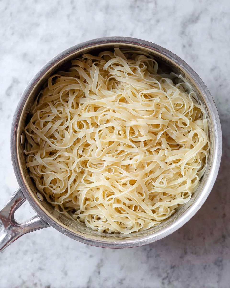 A metal pot filled with a single layer of cooked pale yellow noodles that are thin and flat, tangled lightly and sitting loosely inside the pot. The pot has a shiny silver finish with a round handle visible on the left side. The pot rests on a white marbled surface. photo taken with an iphone --ar 4:5 --v 7