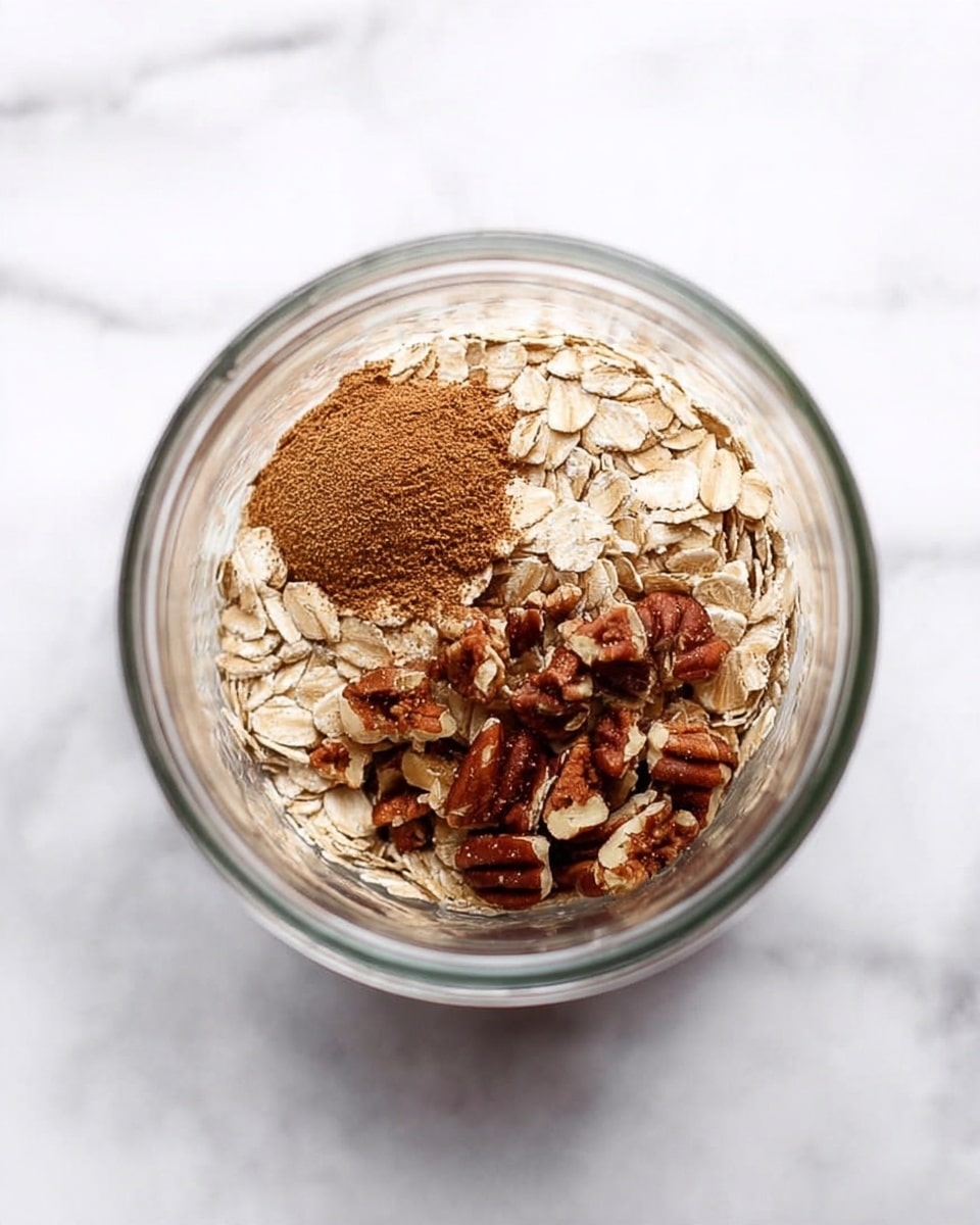 Inside a clear glass jar are three layers sitting on a white marbled surface. The first layer is light beige rolled oats, covering about half the visible space. Next to it, there is a small mound of ground brown cinnamon in the center-left area. On the right side is a layer of chopped pecans, showing a mix of dark brown and tan pieces with rough textures. The jar's round shape frames the ingredients neatly, and soft light highlights the natural colors of the oats, cinnamon, and nuts. Photo taken with an iphone --ar 4:5 --v 7