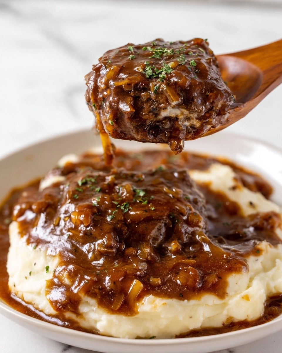 A white plate holds a thick layer of creamy, smooth mashed potatoes with a light white color and soft texture. On top of the mashed potatoes sits a large, thick beef patty covered in a dark brown, glossy gravy mixed with small bits of cooked onions and herbs, giving it a shiny and slightly chunky look. A wooden spoon lifts a generous piece of the beef patty coated in the same rich, dark brown gravy that is dripping slightly, showing the moist and tender texture of the meat. There is a sprinkle of finely chopped green herbs on top of the beef. The dish rests on a white marbled surface, and the photo is taken with an iphone --ar 4:5 --v 7