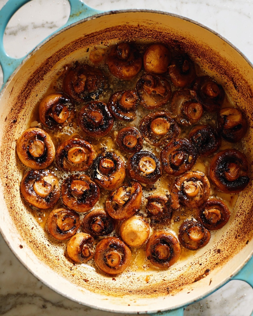 Inside a light blue pot with a white inner surface, there are cooked, soft brown mushrooms with dark grill marks scattered evenly across the bottom. The mushrooms seem to be simmering in a thin layer of butter or oil, which creates tiny bubbles around their edges. The pot's white marbled surface is seen under and around the mushrooms, with some dark browned spots from cooking. photo taken with an iphone --ar 4:5 --v 7