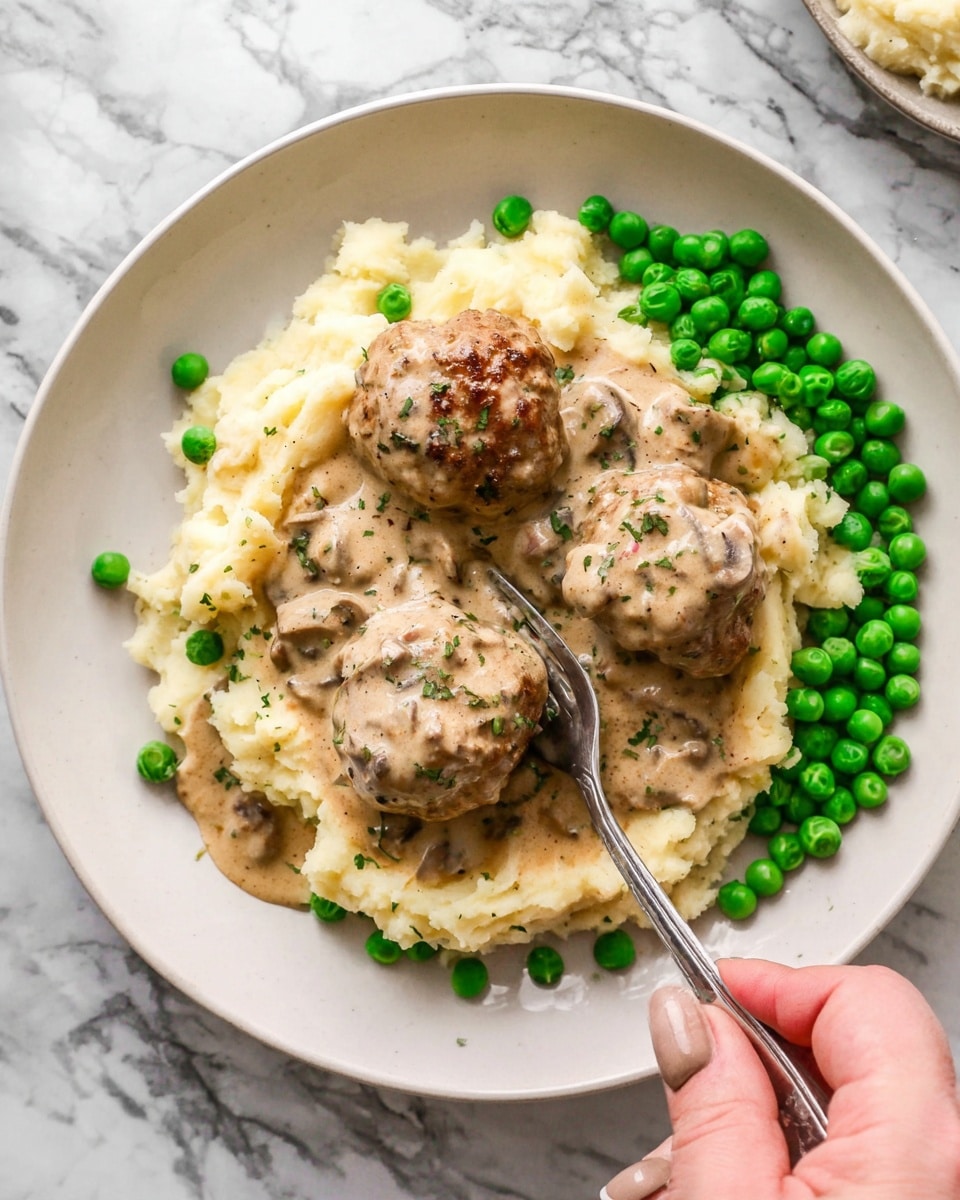 This image shows a white round plate filled with mashed potatoes as the bottom layer, creamy and light beige in color, spreading across most of the plate. On top of the mashed potatoes are three brown meatballs covered with creamy mushroom gravy, giving a smooth texture with small mushroom pieces visible. Around the edges of the plate, there are bright green peas scattered evenly, adding a fresh pop of color. A silver fork is placed in the center of the plate, partially under the gravy, with a woman's hand holding the fork in the bottom right corner. The background features a white marbled surface. Photo taken with an iphone --ar 4:5 --v 7