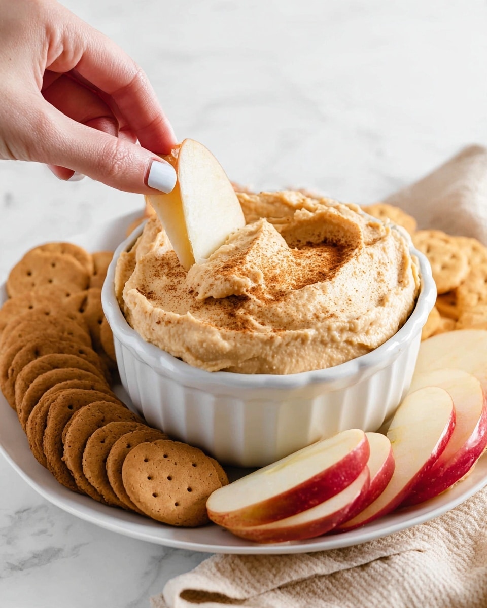 A white ruffled bowl in the center is filled with a thick, light brown dip sprinkled with a darker spice on top. A woman's hand with white nail polish dips a pale apple slice into the dip, lifting it slightly. Around the bowl on a white plate are several pieces of light tan graham crackers, round brown cookies, and sliced red apples with pale yellow inside, all arranged neatly. The background is a white marbled texture. photo taken with an iphone --ar 4:5 --v 7