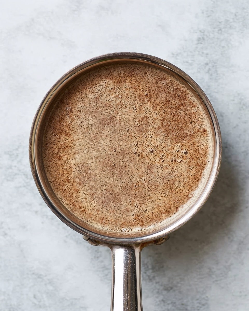 A top view of a shiny metal pan filled with a light brown liquid that has tiny bubbles on the surface and darker brown specks swirling softly throughout the mixture. The pan handle extends from the bottom of the frame, resting on a white marbled surface. The colors are warm and earthy, showing the contrast between the frothy liquid and the smooth metal pan rim photo taken with an iphone --ar 4:5 --v 7
