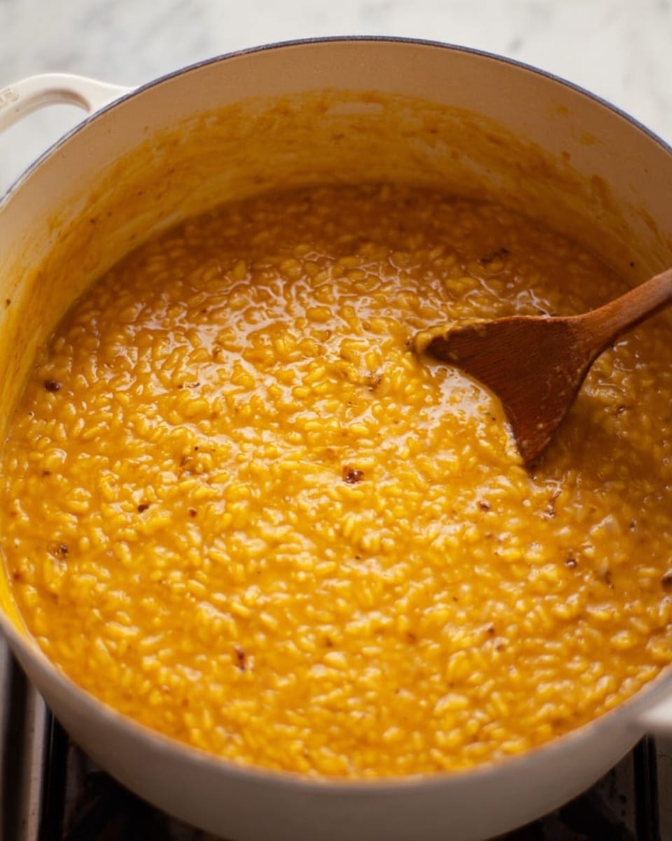 This image shows a close-up of a thick, creamy orange risotto being stirred inside a large white pot. The risotto has visible grains that create a textured surface and small darker spots spread throughout. On the right side, a wooden spoon is partially submerged in the risotto, blending with the smooth and slightly shiny texture. The pot sits on a stove, and the background is a white marbled surface. photo taken with an iphone --ar 4:5 --v 7